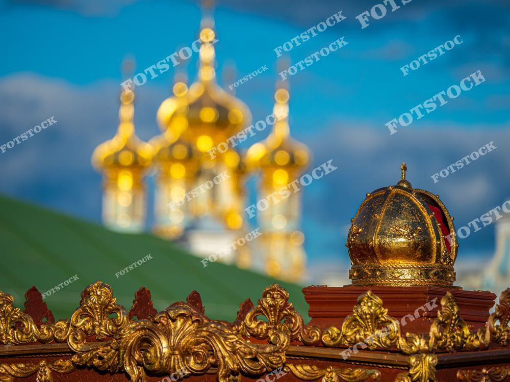 На фотографии изображены золотые купола Екатерининского дворца в Царском Селе, одном из выдающихся памятников архитектуры Санкт-Петербурга. На переднем плане — декоративная золотая корона, являющаяся частью оформления дворцовой территории. Солнечные блики подчеркивают роскошь и великолепие барочных элементов. Фотография передает величие дворцово-паркового ансамбля.